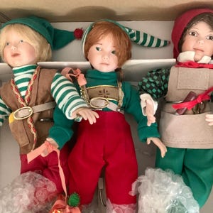 May include: Three porcelain dolls dressed as elves. The elf on the left is wearing a green and brown outfit with a brown belt and a green hat. The elf in the middle is wearing a red and green outfit with a green hat. The elf on the right is wearing a green and brown outfit with a brown apron and a red hat.