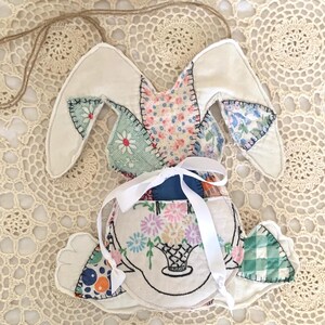 May include: A handmade Easter bunny decoration. The bunny is made of fabric scraps in various floral and patterned designs. It has long ears, a white ribbon, and a basket of flowers embroidered on the front. A jute string is attached for hanging.