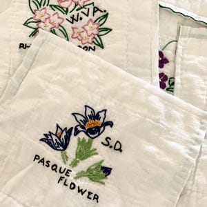 May include: White embroidered fabric squares with floral designs. One square features pink flowers with green leaves and the text "W.V.A." and "RH...ON". Another square displays blue and yellow flowers with the text "PASQUE FLOWER" and "S.D."