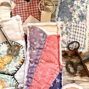 May include: Close-up of several handmade fabric keychains with metal key rings. The keychains feature patchwork designs in various colors and patterns, including floral, plaid, and solid fabrics. Some keychains have a ribbon loop. Antique keys are also visible.