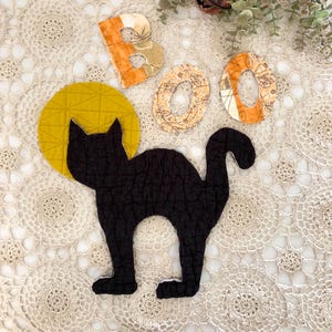 May include: A quilted black cat silhouette positioned in front of a mustard yellow circular moon shape, accompanied by three fabric letters spelling BOO in orange and cream patterned textiles, featuring a textured, stitched surface finish.