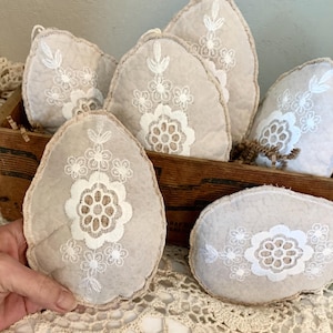 May include: A collection of beige, egg-shaped decorative items with white floral embroidery. The items are displayed in a wooden crate and on a lace surface. The embroidery features a central flower design with surrounding smaller floral accents.