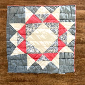 May include: Square quilted textile with a starburst pattern. The design features a central white diamond shape, surrounded by red and white triangles, and blue floral print triangles. The border is a muted blue-gray color.