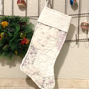 May include: A white quilted Christmas stocking with a floral pattern and a white cuff. The stocking is hanging from a metal hook on a white wall.