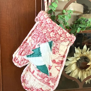 May include: A quilted oven mitt with a red and white floral pattern and a green and white tree applique. The mitt is hanging from a dark brown cabinet door. The overall theme is kitchen decor.