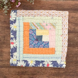 May include: A square, handmade quilt with a log cabin design. The center features a patchwork of orange, blue, pink, and yellow fabrics. The quilt is bordered with a blue floral fabric and a patterned edge. The quilt is placed on a wooden surface.