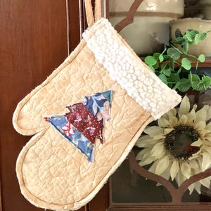 May include: Tan quilted oven mitt with a cream-colored sherpa cuff and a fabric Christmas tree applique. The tree is made of red, blue, and floral patterned fabric. A loop is attached for hanging.