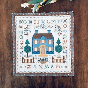 May include: A square fabric sampler featuring a cross-stitch design of a blue house with a brown roof, surrounded by a border of the alphabet in blue and brown. The design includes two green trees, a fence, a flower basket, and birds.