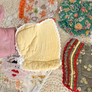 May include: A collection of vintage baby bonnets in various colors and patterns. The bonnets feature floral prints, ruffles, and lace trim. One bonnet is yellow with a quilted texture, and another has a face design.