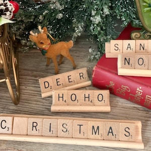 May include: Wooden letter tiles spelling out the words "DEER", "HO HO", "CHRISTMAS", and "SAN", "NO". The tiles are arranged on a wooden surface with a small, brown, plush reindeer figurine in the background.