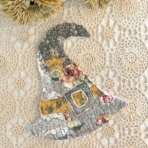 May include: A quilted witch hat with a patchwork design. The hat features floral, star, and geometric patterns in shades of gray, yellow, and burgundy. The hat is likely a decorative item for Halloween or fall.