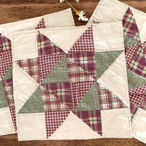 May include: A set of square fabric coasters featuring a star pattern constructed from patchwork pieces. The design incorporates cream-colored fabric alongside plaid and checkered patterns in shades of deep red, forest green, and tan.