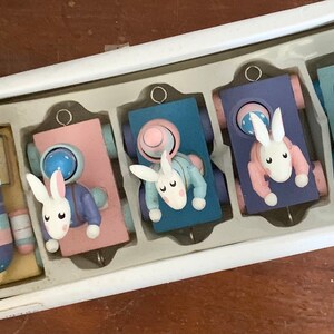May include: A set of five wooden train cars decorated with pastel colors and bunny faces. Each car has a different color and features a bunny wearing a different colored outfit. The train cars are packaged in a white box.