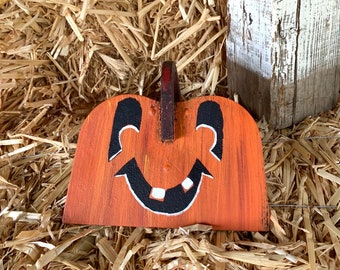 May include: A hand-painted wooden pumpkin with a smiling face. The pumpkin is orange with black eyes, a black mouth, and white teeth. The pumpkin is sitting on a bed of straw.