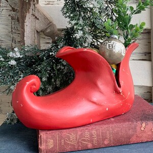 May include: A red ceramic elf shoe with a green felt lining. The shoe is filled with vintage Christmas ornaments, including a silver ornament with the letter "S" on it.