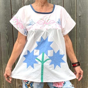 May include: A white cotton tunic top with blue and green floral embroidery. The top has a round neckline and short sleeves. The embroidery features three blue flowers with green stems and leaves.