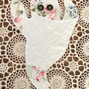 May include: A handmade ghost-shaped quilted item with floral fabric accents. The ghost features a white quilted body, floral fabric arms, and dark green button eyes. The background is a cream-colored lace doily on a wooden surface.