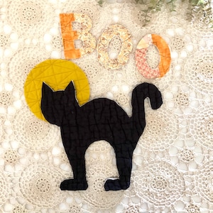 May include: A quilted black cat silhouette positioned in front of a circular yellow quilted moon. Above the cat are three fabric letters spelling the word BOO, with the B and O in orange floral patterns and the final O in a pink floral print.