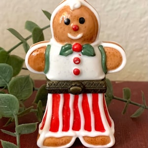 May include: A porcelain gingerbread man figurine with a white shirt, green bow tie, red and white striped pants, and a brown belt. The figurine is a trinket box with a hinged lid.