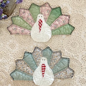 May include: Two handmade turkey-shaped fabric decorations. Each turkey has a white body with a red and white checkered wattle. The fan-shaped tail feathers are a mix of green, pink, and blue floral patterns, outlined with black stitching. The turkeys are on a cream-colored lace surface.