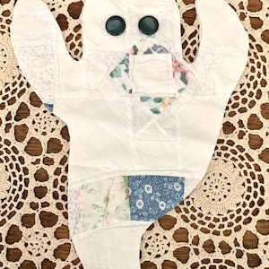 May include: A handmade ghost-shaped quilt with a patchwork design. The ghost is primarily white, with patches of floral fabric in shades of pink, blue, and green. Two dark green buttons serve as eyes. A unique, whimsical Halloween decoration.
