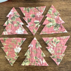 May include: Seven fabric Christmas tree ornaments. Each tree is cut from a floral pattern fabric with a pink background and white and green floral details. The trees are arranged on a wooden surface.