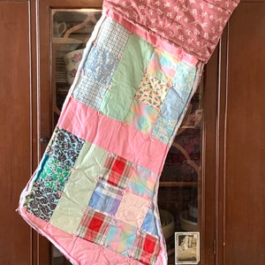 May include: A quilted Christmas stocking featuring a patchwork design of various fabric squares in shades of pink, blue, green, and red. The cuff is made of a solid pink fabric with a white floral pattern, and the entire piece is tied with small knots.