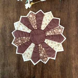 May include: A circular quilted fabric coaster featuring a dahlia-like pattern with alternating segments of solid mauve fabric and cream fabric printed with a brown floral motif, finished with a central solid mauve circle and white edge binding.