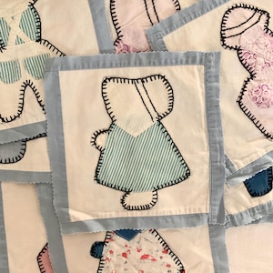 May include: A collection of handmade fabric squares featuring appliqued figures. Each square has a white background with a light blue border. The figures are stitched with black thread and include dresses and overalls with patterned fabric.