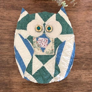 May include: A quilted fabric owl ornament featuring a patchwork design in shades of teal, white, and light blue. The owl has two round cream-colored fabric eye patches with gold and teal button centers and a small floral patterned fabric beak.