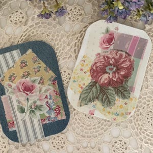 May include: Two decorative, rectangular patches with floral designs. One patch is denim blue with a pink rose and yellow floral accents. The other is white with a large burgundy flower and pink and yellow floral accents.