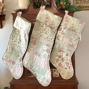 May include: Three quilted Christmas stockings with a floral pattern in shades of pink, green, and white. The stockings are hung on a wooden mantel.