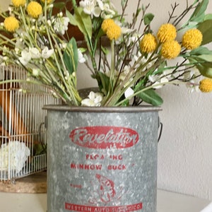 May include: A vintage galvanized metal minnow bucket with the words "Revelation Floating Minnow Buck" and "Western Auto Supply Co." printed in red. The bucket is filled with a bouquet of white and yellow flowers.
