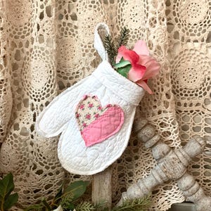 May include: White quilted oven mitt with a pink and floral heart applique. The mitt is filled with pink flowers and is displayed on a wooden post.