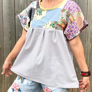 May include: A white and gray striped shirt with a patchwork design on the yoke. The patchwork is made of various fabrics in different colors and patterns. The shirt has short sleeves and a round neckline.
