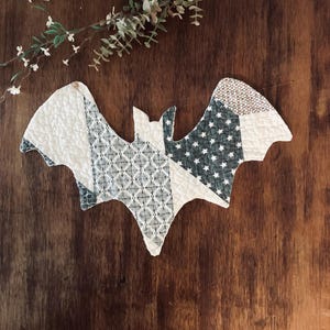 May include: A quilted fabric bat ornament featuring a patchwork design of white, dark green, and grey fabrics. The patterns include small white stars on a dark green background, a geometric diamond print, and solid white textured sections.