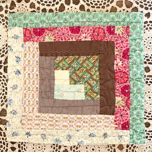 May include: A square, handmade quilt with a log cabin design. The center features a brown square surrounded by patterned fabrics in shades of pink, green, and cream. The quilt is likely made of cotton and has a vintage aesthetic.
