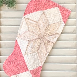 May include: A pink and white patchwork Christmas stocking with a large white star in the center. The stocking is hanging on a white wooden shutter.