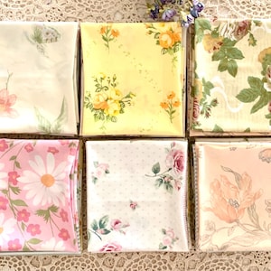 May include: Six folded fabric squares with floral patterns. The patterns include daisies, roses, and other flowers in shades of pink, yellow, and cream. The fabrics are arranged on a lace surface.