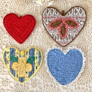 May include: Four heart-shaped quilted pot holders in red, brown, blue, and yellow. The red holder has a quilted design. The brown holder has a pink bow design. The blue holder has a white lace trim. The yellow holder has a flower design.