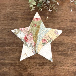 May include: A star-shaped fabric ornament featuring a quilted texture and a patchwork design. The surface displays various floral patterns in shades of pink, green, yellow, and cream, with visible stitching throughout the entire star-shaped piece.