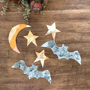 May include: A collection of Halloween-themed fabric cutouts, including a crescent moon, stars, and bats. The moon is orange and yellow, while the stars are yellow and white. The bats are gray with a floral pattern. The items are arranged on a wooden surface.