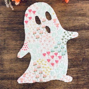May include: A ghost-shaped quilted fabric cutout featuring a white background with a repeating pattern of small pink, yellow, and light green flowers. The ghost has two oval-shaped eye cutouts and one oval-shaped mouth cutout, with visible stitching.