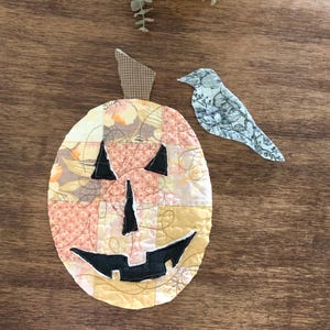 May include: A quilted fabric pumpkin decoration featuring a patchwork design of various floral and geometric patterns in shades of yellow, orange, and cream. It has a brown checkered stem and black fabric eyes, nose, and a wide, jagged mouth.