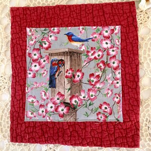 May include: A square, quilted fabric piece with a central image of bluebirds and a birdhouse surrounded by pink flowers. The central image is set against a gray background and framed by a red, textured border.