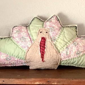 May include: A handmade turkey-shaped pillow with a beige body and a red wattle. The fan-shaped tail features green and pink floral patterned fabric, outlined with black stitching. A small pumpkin decoration is visible on the left.