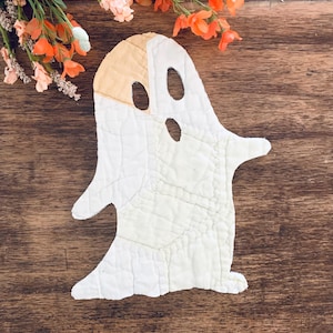 May include: A white quilted ghost-shaped fabric cutout with a tan patch on the upper left side. The ghost features two oval eye cutouts and one oval mouth cutout, with visible stitching throughout the quilted surface and a textured appearance.