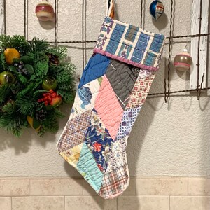 May include: A patchwork Christmas stocking with a blue, pink, and white color scheme. The stocking is hanging on a wire fence.