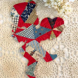May include: A handmade patchwork figure in a whimsical design. The figure is primarily red, with sections of patterned fabric in blue, white, and other colors. The figure is placed on a cream-colored, lacy surface, with white flowers in the background.