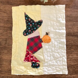 May include: A rectangular fabric wall hanging featuring a quilted background split between pale yellow and white. A central witch figure is appliqued using various fabric scraps, including a black floral hat, red plaid bodice, and a red bandana skirt.